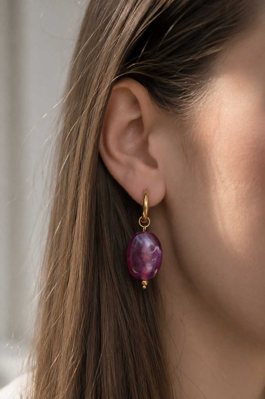 Goldene Creolen mit lila-violetter Marmorperle am Ohr getragen, vergoldete Huggie-Ohrringe mit ovalem Achat-Anhänger und goldenem Kugeldetail zu braunem Haar