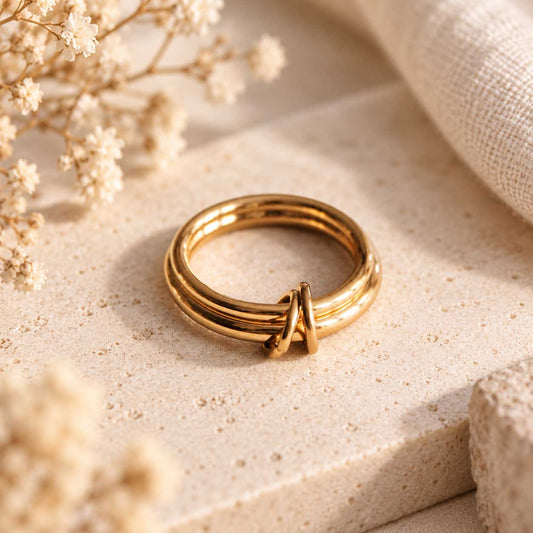 Double Loop goldener Ring – vergoldeter Edelstahlring mit zwei parallelen Bändern und Knotendetail in der Mitte, auf Travertin-Stein mit Schleierkraut, warmes Licht