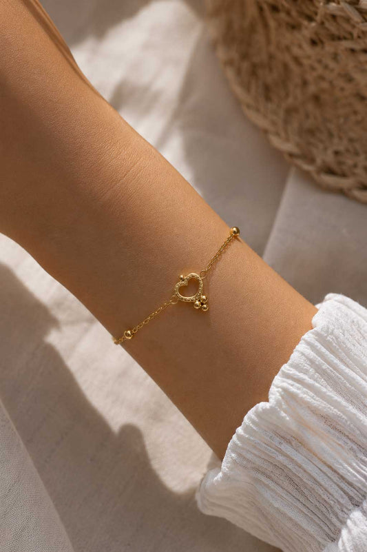 Golden Glow Armband am Handgelenk – goldenes Herzarmband von Saltyblush getragen, offener Herzcharm mit Kugeldetails zu weißem Strickoberteil im Sonnenlicht