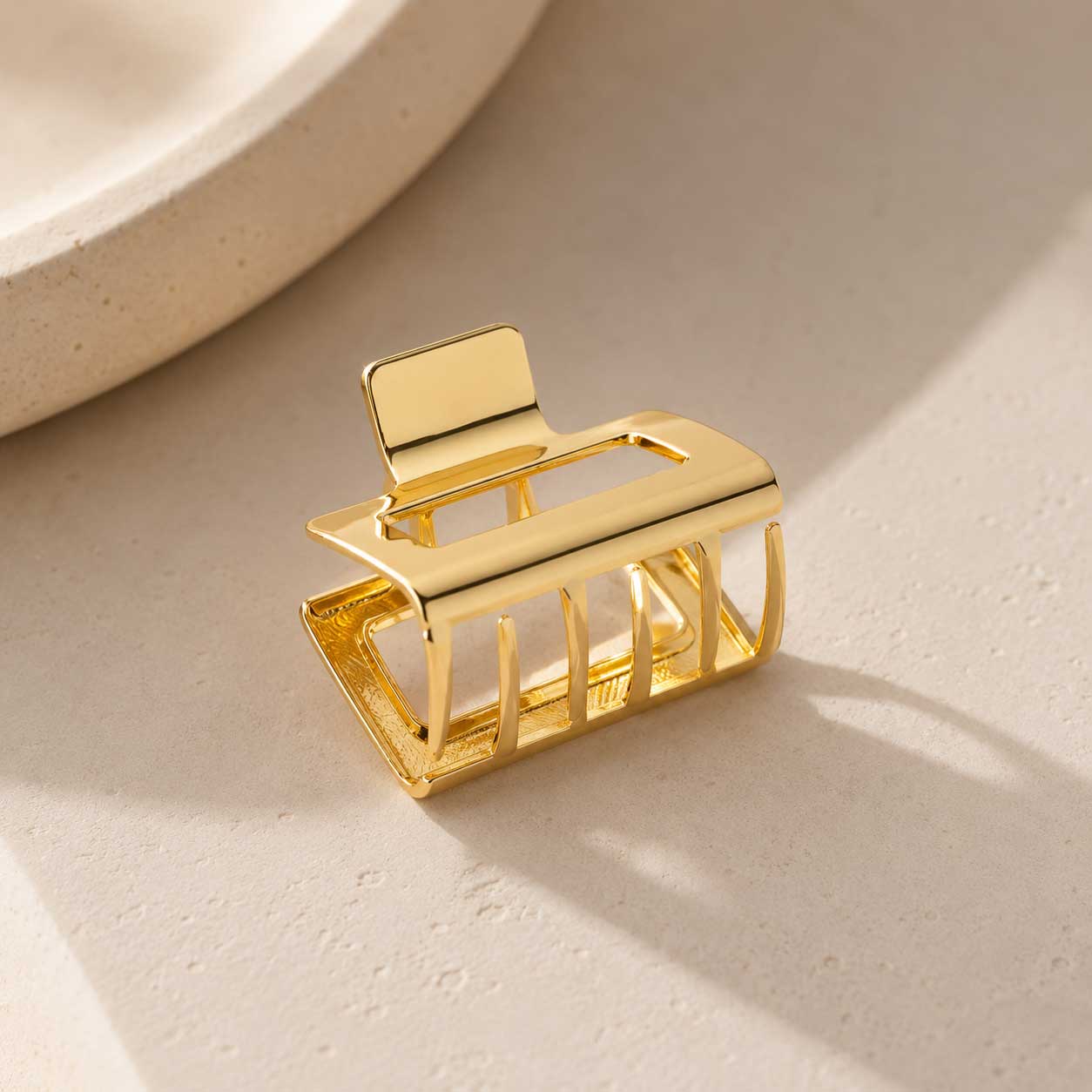Goldene rechteckige Haarklammer Seitenansicht auf cremefarbenem Stein im Sonnenlicht, vergoldete Metall-Haarspange mit offenem Gitter-Rechteck-Design und Schattenspiel