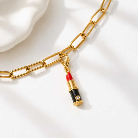 Goldener Lippenstift-Charm an vergoldeter Paperclip-Kette befestigt, roter Emaille-Charm mit schwarzem Gehäuse und Zirkonia-Stein auf weißem Seidenstoff