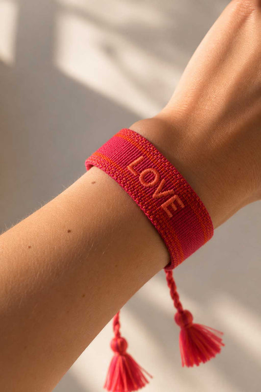 Rotes LOVE Armband am Handgelenk getragen, gewebtes Stoffarmband mit Quasten im Sonnenlicht