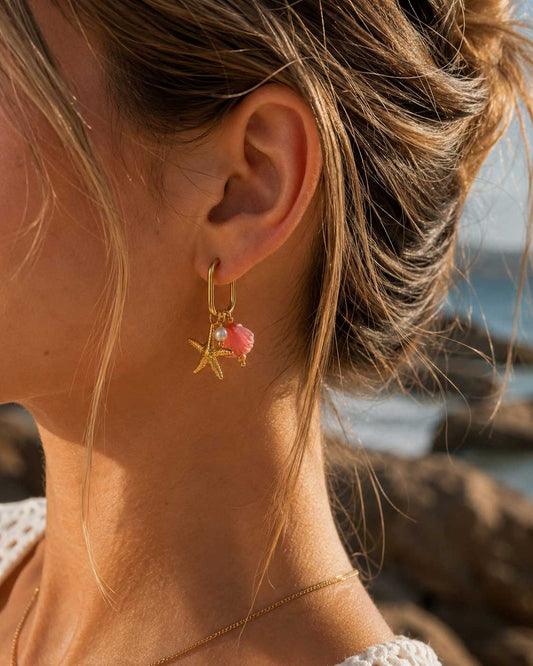 Frau trägt pearl and sea Ohrringe am Ohr – goldene Creolen mit Seestern- und Muschel-Charm