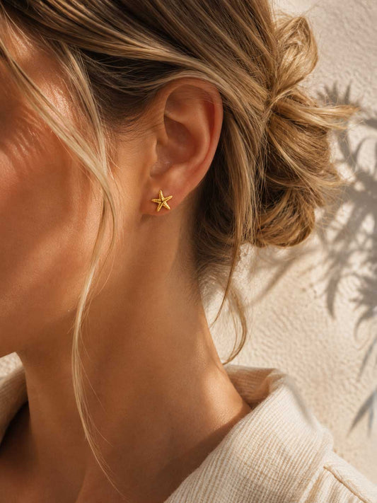 Sea Spirit Ohrringe Lifestyle – goldene Seestern Stecker von Saltyblush am Ohr einer Frau mit blondem Haar, sommerlicher Strandlook mit Palmenschatten im Hintergrund