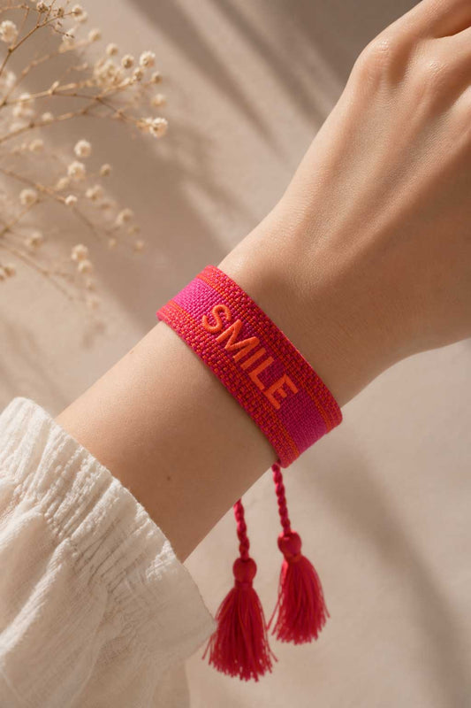 Pinkes SMILE Armband mit roten Quasten am Handgelenk getragen, weißes Kleid, Trockenblumen im Hintergrund – Saltyblush