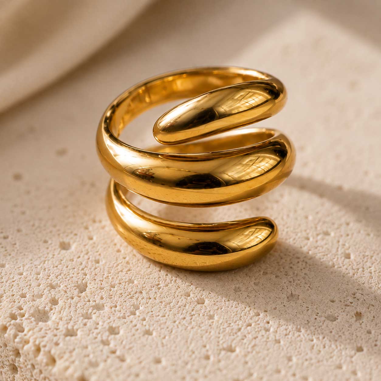 Nahaufnahme des Soft Curve goldenen Rings aus Edelstahl mit dreilagiger Spiralform auf cremefarbenem Travertin – Saltyblush
