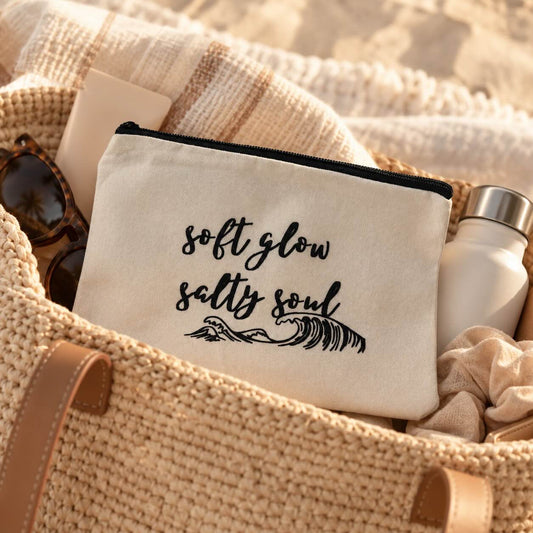 Die beige soft glow salty soul Kosmetiktasche mit schwarzem Wellenmotiv in einer geflochtenen Strandtasche, neben Sonnenbrille, Handtuch und Trinkflasche.