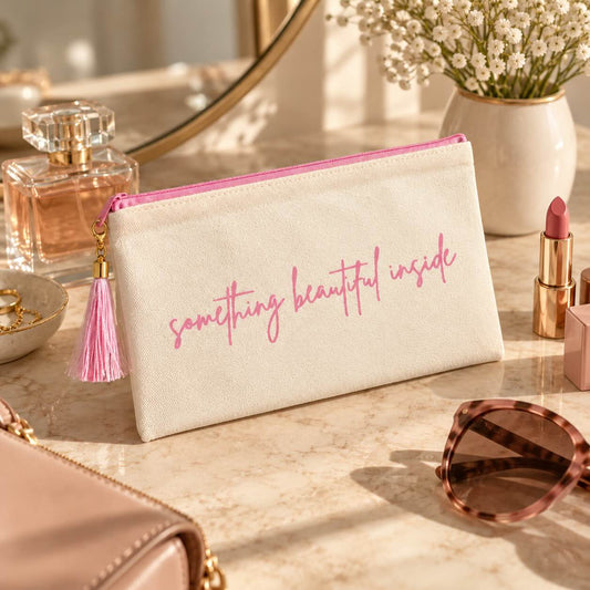 Beige something beautiful inside Kosmetiktasche mit rosa Schriftzug und Quaste vor einem runden Spiegel, neben Parfum, Lippenstift, Sonnenbrille und weißen Blumen.