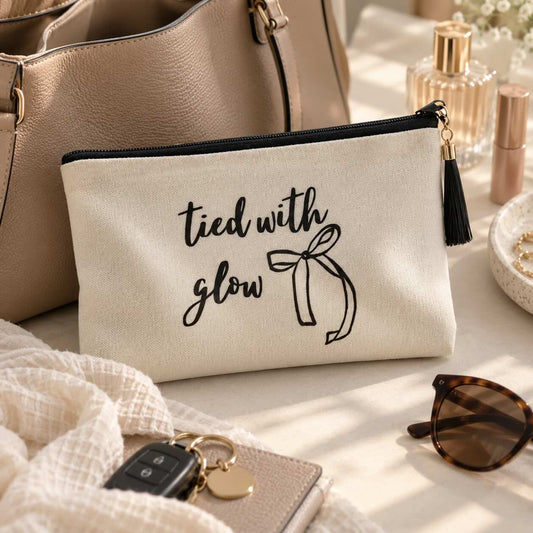 Beige tied with glow Kosmetiktasche mit Schleifenmotiv und schwarzer Quaste vor einer Handtasche, neben Sonnenbrille, Schlüssel und Kosmetikprodukten.