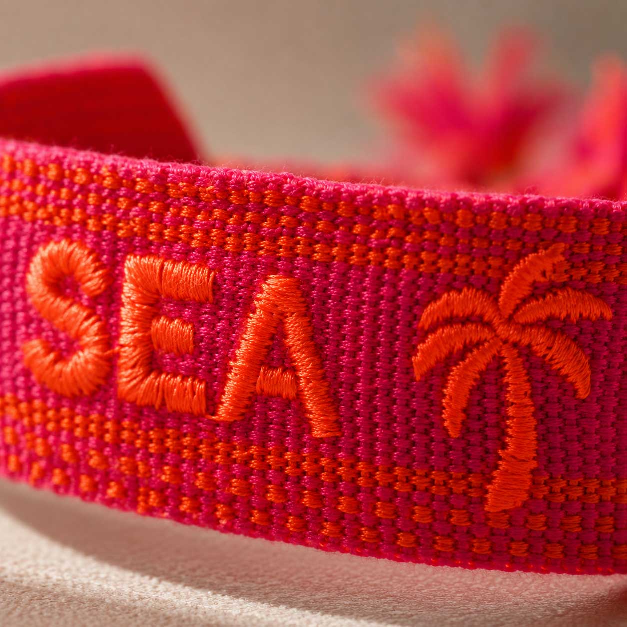 Nahaufnahme der orangen SEA Stickerei und Palmen-Motiv auf pinkrot gewebtem Textilarmband – Saltyblush