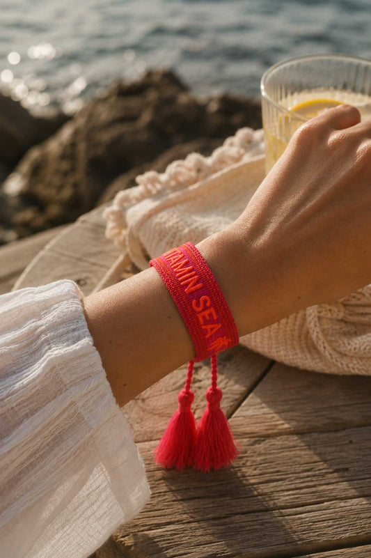 Rotes Vitamin Sea Armband mit roten Quasten am Handgelenk getragen, Meeresblick und Holzsteg im Hintergrund – Saltyblush