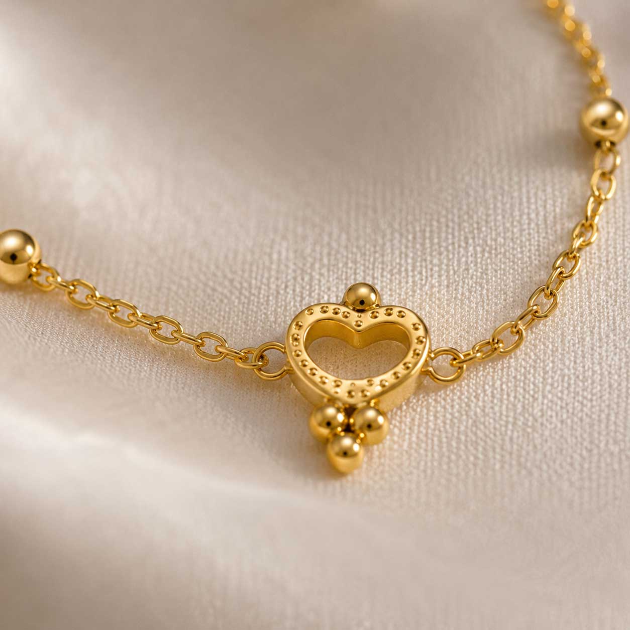 Golden Glow Armband Flatlay – goldenes Herzarmband von Saltyblush, offener Herzcharm mit 3 Kugelanhängern an Satellitenkette, auf weißem Stoff mit Trockenblumen
