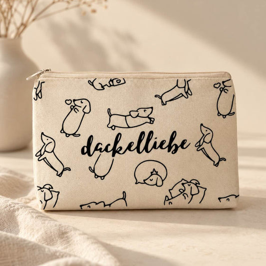 Beige Dackelliebe Kosmetiktasche mit Reißverschluss, schwarzen Dackelzeichnungen in verschiedenen Posen und mittigem Schriftzug „dackelliebe“ auf hellem Tisch.
