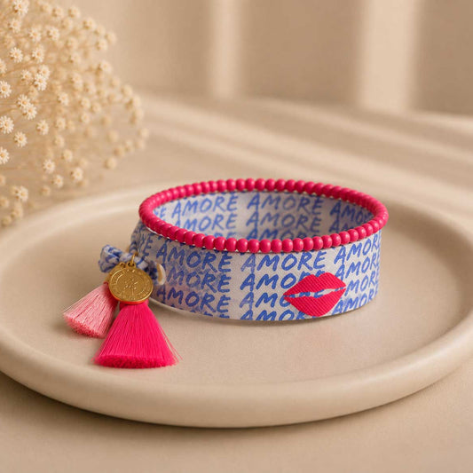Elastisches Armband-Set Amore Kiss mit weiß-blauem Amore-Schriftzug-Band, pinkem Perlenarmband, pinker Quaste und goldenem Anhänger auf Keramikteller – Saltyblush

