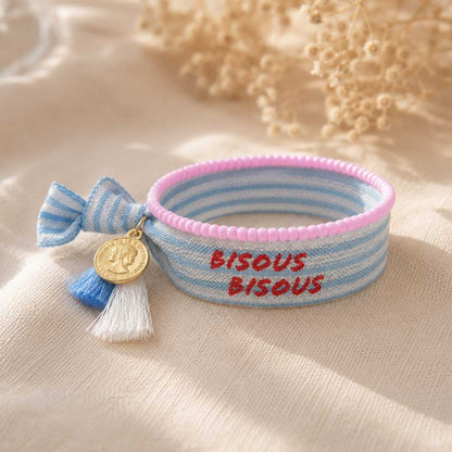 Elastisches Armband-Set Bisous Bisous mit hellblau-weißem Streifenband, rotem „Bisous Bisous"-Schriftzug, blau-weißer Quaste, goldenem Münzanhänger und rosa Perlenarmband auf cremefarbenem Stoff – Saltyblush
