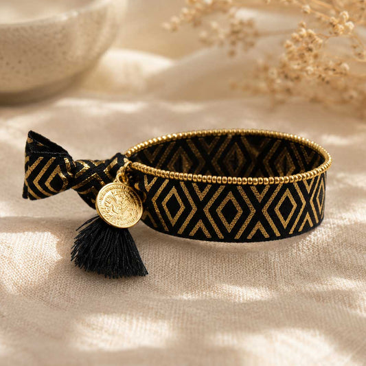 Elastisches Armband-Set Black & Gold mit schwarzem Rautenmuster-Band, schwarzer Quaste und goldenem Münzanhänger sowie goldenem Perlenarmband auf cremefarbenem Leinenstoff – Saltyblush
