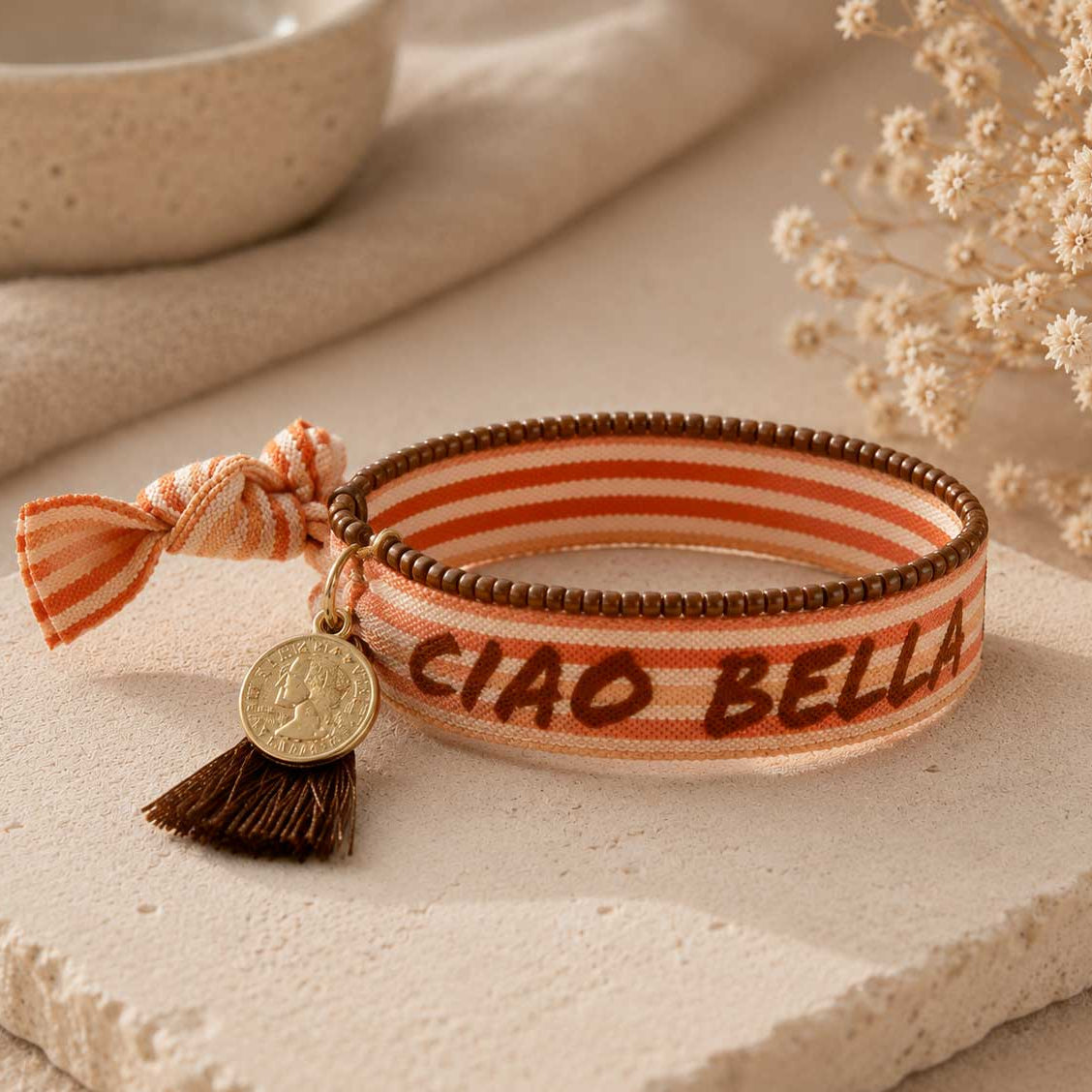 Elastisches Armband-Set Ciao Bella mit orangefarbenem Streifen-Schriftzugband, brauner Quaste und goldenem Münzanhänger auf Steinuntergrund – Saltyblush

