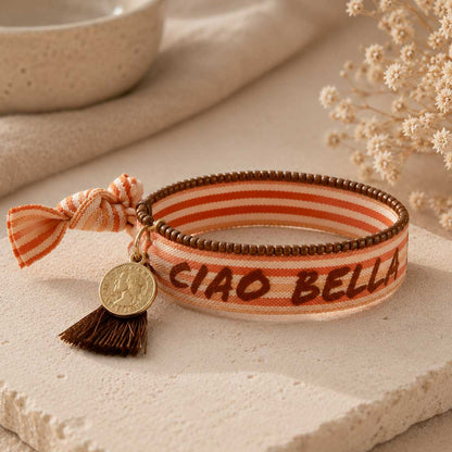 Elastisches Armband-Set Ciao Bella mit orangefarbenem Streifen-Schriftzugband, brauner Quaste und goldenem Münzanhänger auf Steinuntergrund – Saltyblush
