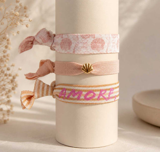 Elastisches 3er Armband-Set Soft Amore mit rosa Kuss-Print-Band, pfirsichfarbenem Band mit goldenem Muschelanhänger und korallefarbenem „Amore"-Streifenband auf cremefarbener Vase – Saltyblush
