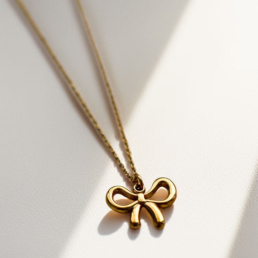 Goldene Halskette Bow Glow mit filigranem Schleifen-Anhänger auf cremefarbenem Hintergrund – Edelstahl, Saltyblush
