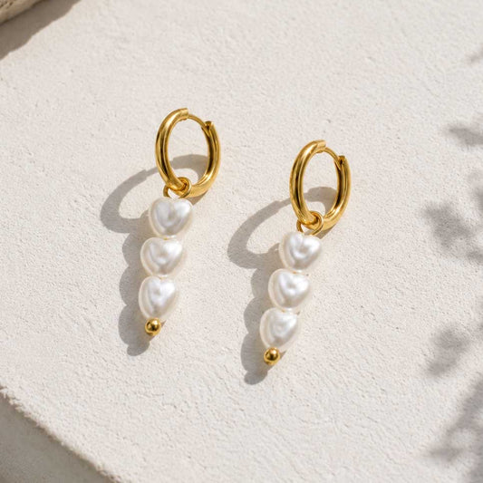 Goldene Creolen Amora mit drei herzförmigen weißen Perlen-Anhängern auf Travertinstein mit Sonnenschatten – Edelstahl, Saltyblush
