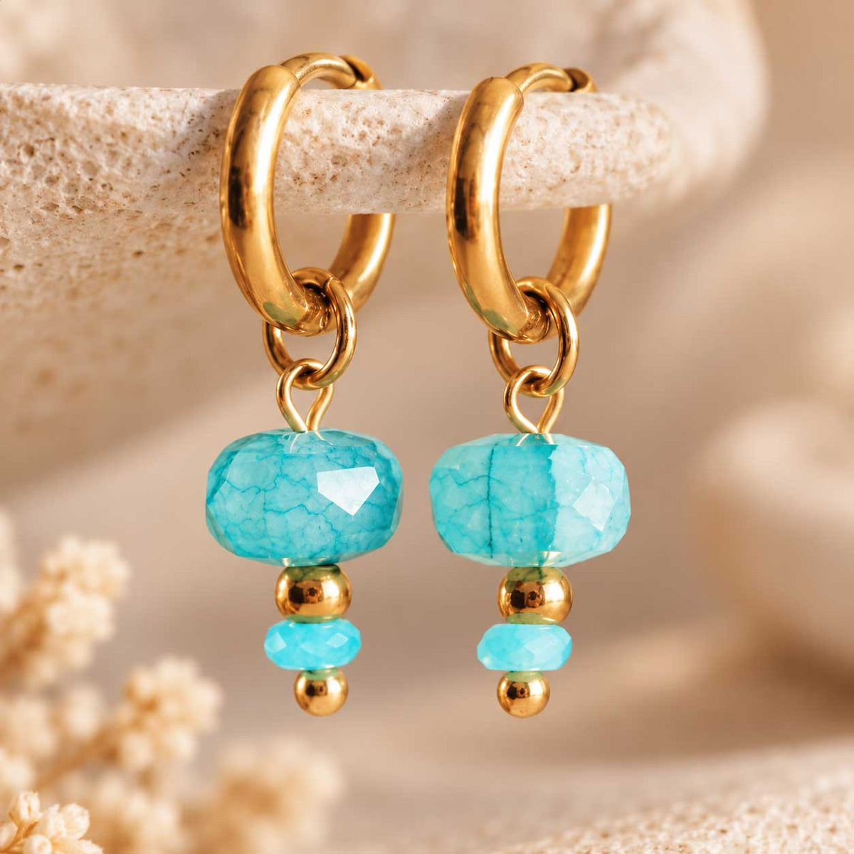 Detailaufnahme einer goldenen Creole aus Edelstahl mit facettierter Optik und auffälligem aqua-blauem Anhänger in tropfenähnlicher Form
