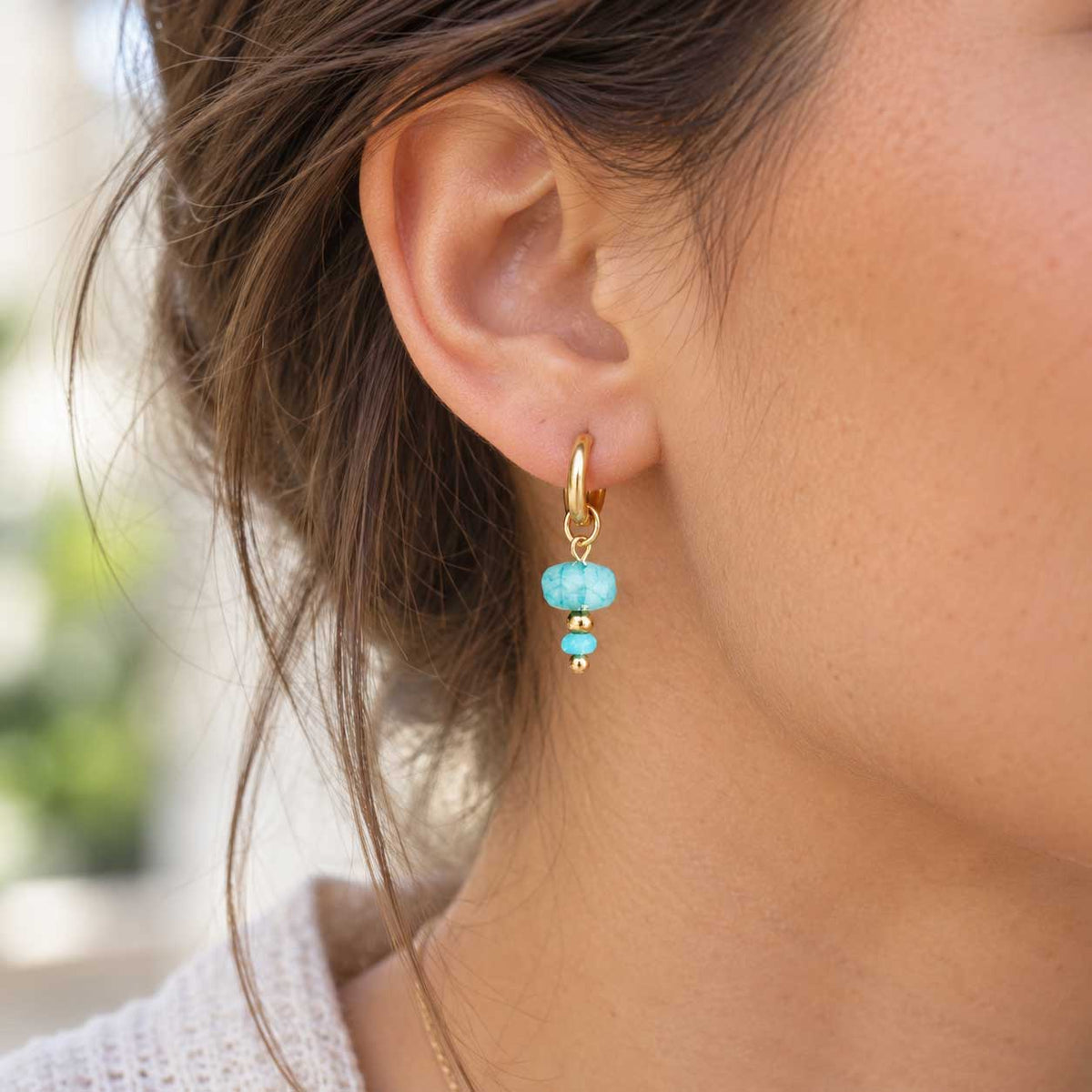 Goldene Creolen aus Edelstahl am Ohr getragen, mit facettierter Oberfläche und aqua-blauem Drop-Charm als frischer Akzent für Alltag und besondere Looks
[AI]