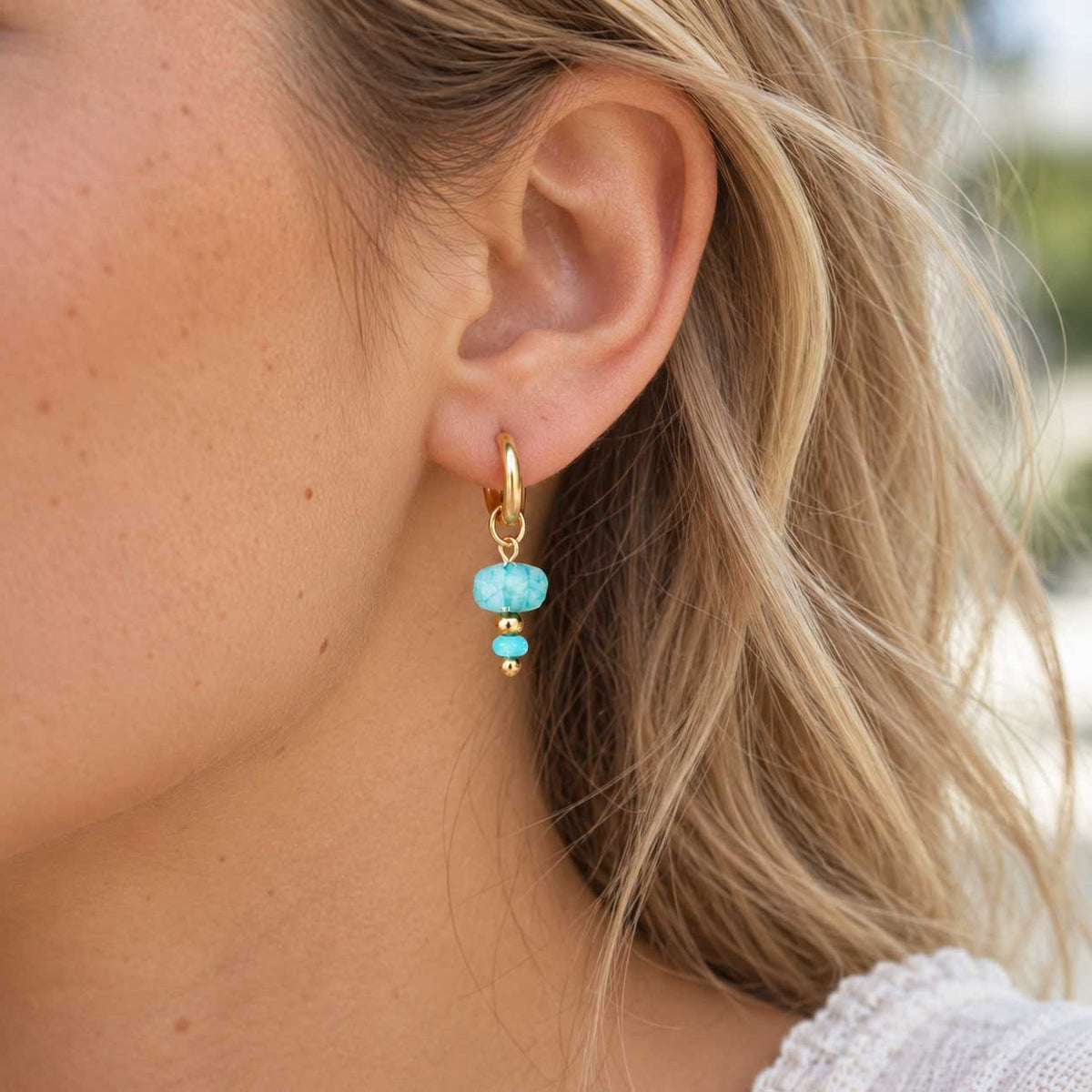 Goldene Creolen aus Edelstahl am Ohr getragen, mit facettierter Oberfläche und aqua-blauem Drop-Charm als frischer Akzent für Alltag und besondere Looks
[AI]