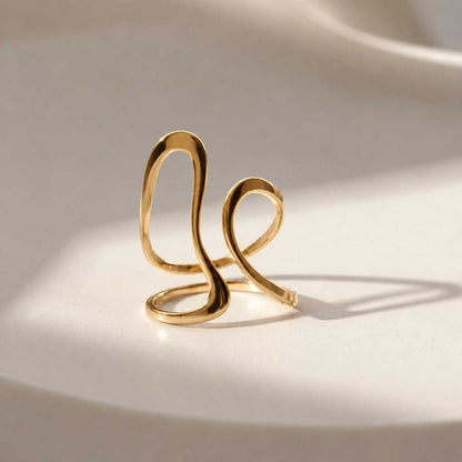 Aura Flow goldener Ring aus Edelstahl mit geschwungener Wellenform auf cremefarbenem Seidenstoff mit Lichtreflexen – Saltyblush
