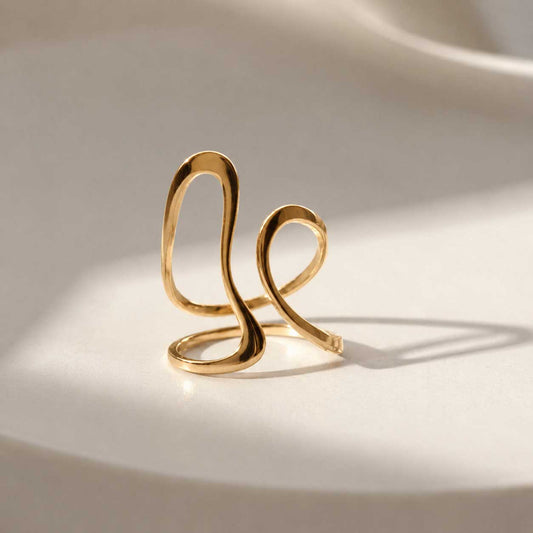 Aura Flow goldener Ring aus Edelstahl mit geschwungener Wellenform auf cremefarbenem Seidenstoff mit Lichtreflexen – Saltyblush
