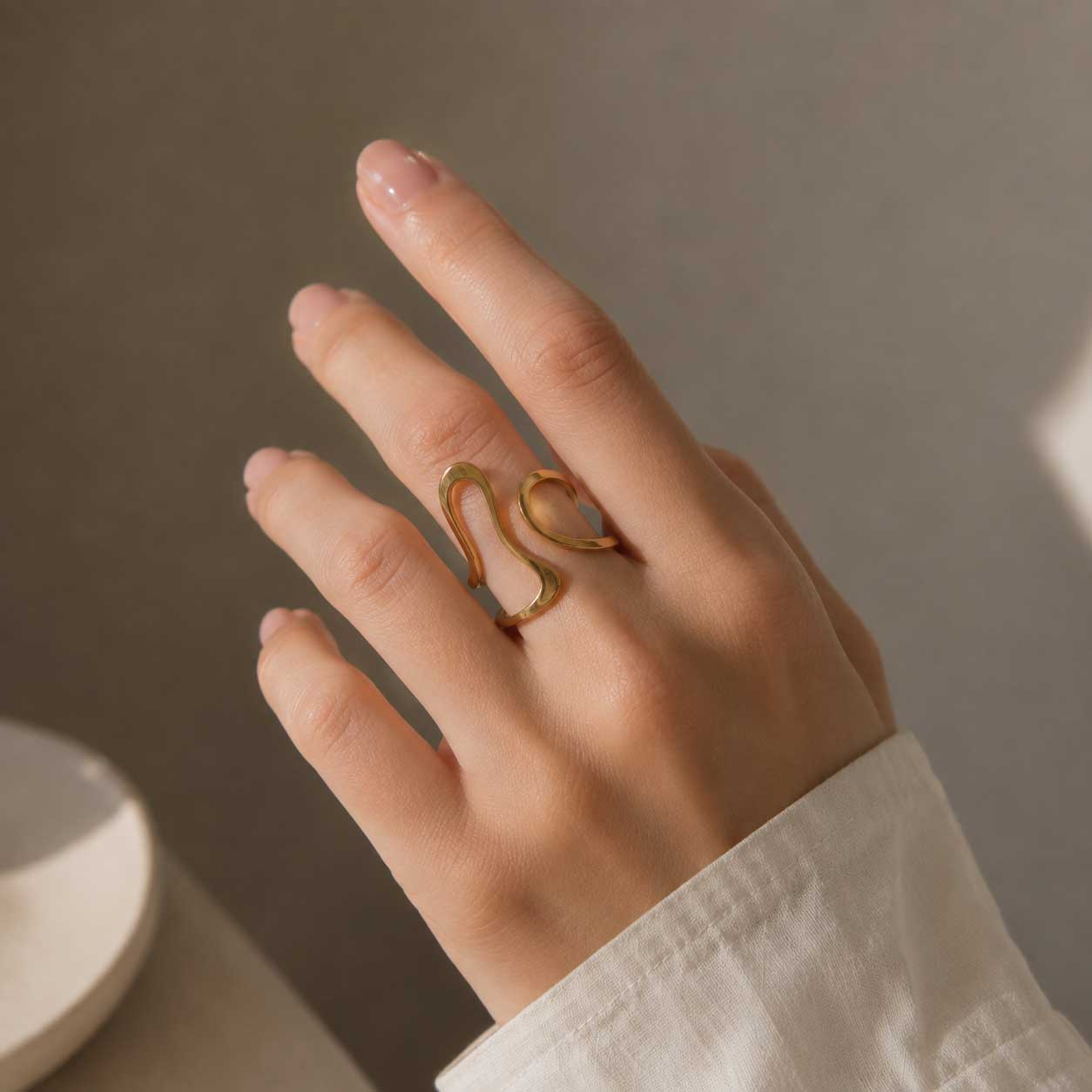 Hand einer Frau mit weißem Hemd trägt den Aura Flow goldenen Ring mit geschwungener Wellenform am Zeigefinger – Saltyblush
