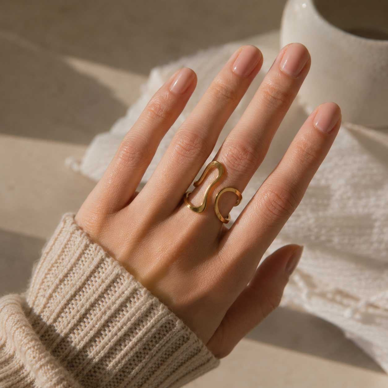 Hand einer Frau mit cremefarbenem Strickpullover trägt den Aura Flow goldenen Ring mit Wellendesign am Mittelfinger – Saltyblush
