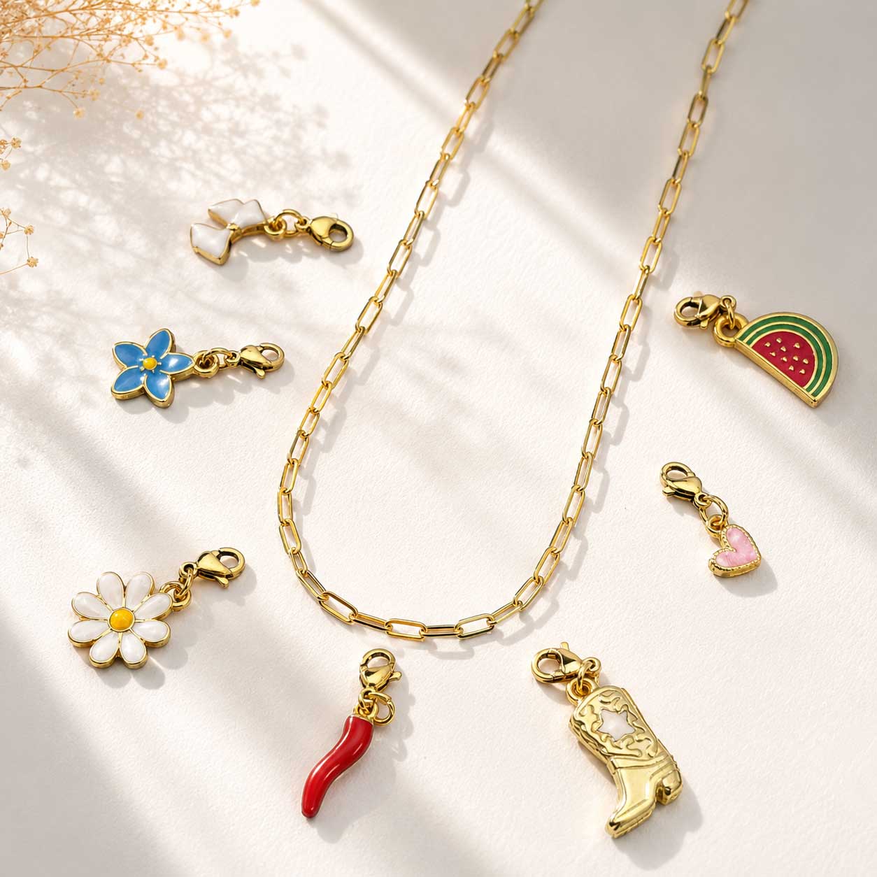 Goldene Paperclip-Halskette als Charm-Basis mit bunten Emaille-Charms – Gänseblümchen, blaue Blume, Schleife, Chili, Cowboystiefel, Wassermelone und Herz auf weißem Hintergrund
