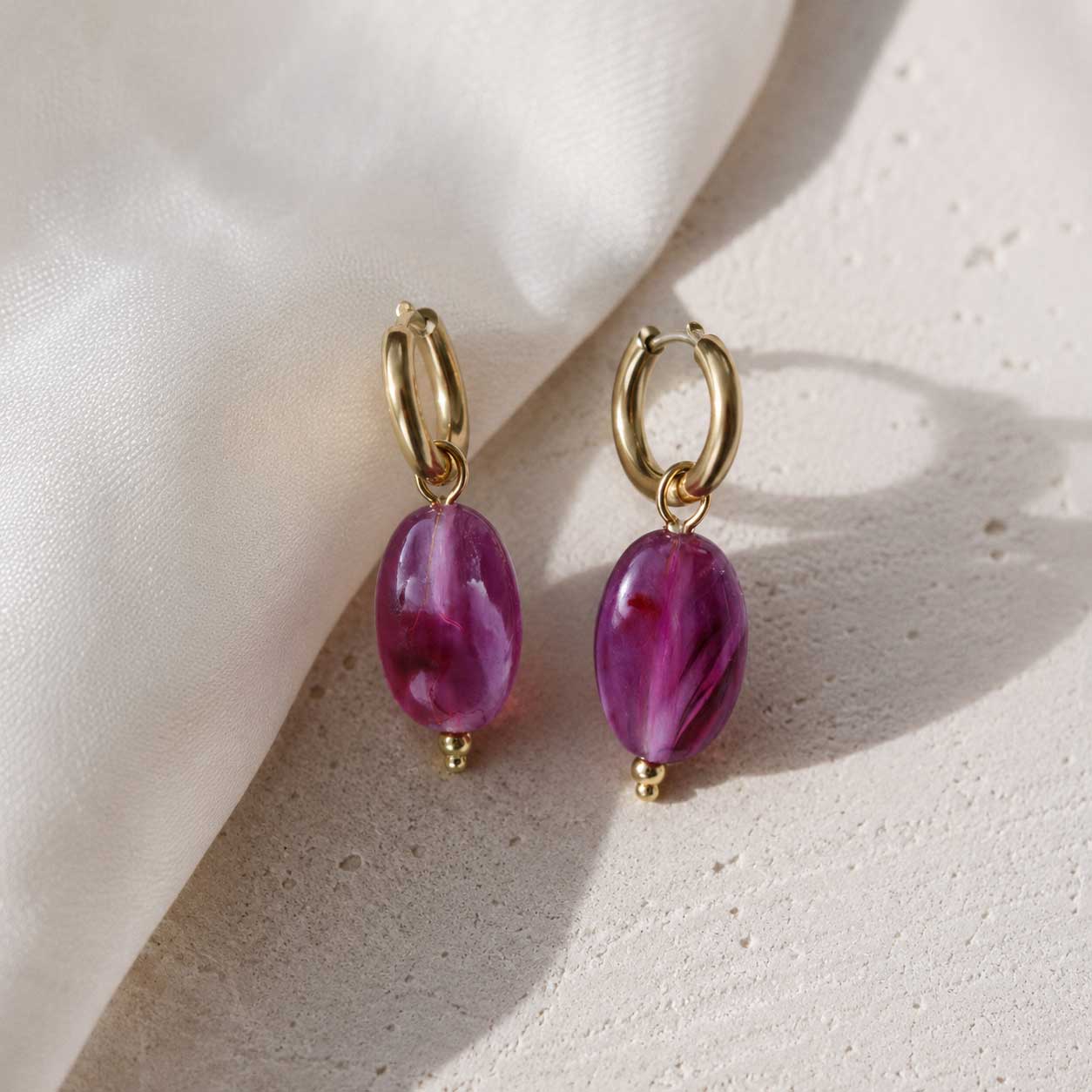 Goldene Creolen mit lila-violetten Marmorperlen auf cremefarbenem Stein mit Seidenstoff im Sonnenlicht, vergoldete Edelstahl-Huggie-Ohrringe mit ovalen Achat-Anhängern
