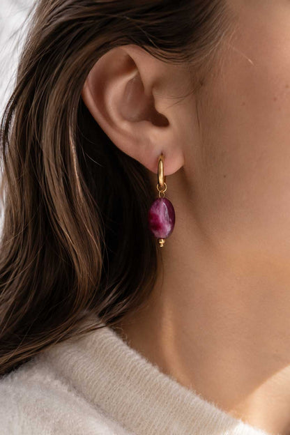 Goldene Creolen mit lila-violettem Marmorstein-Anhänger am Ohr getragen, vergoldete Huggie-Ohrringe mit ovalem Achat-Charm zu weißem Strickoberteil
