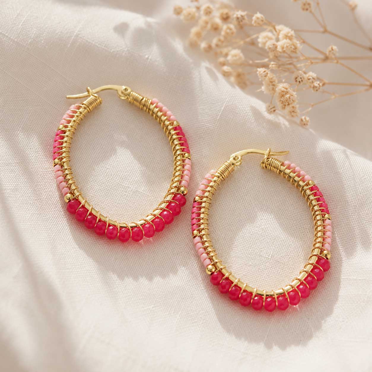 Goldene Creolen mit pinken und hellrosa Perlen auf weißem Leinenstoff mit Schleierkraut, ovale Kristall-Ohrringe mit Farbverlauf in Pink-Rosa
