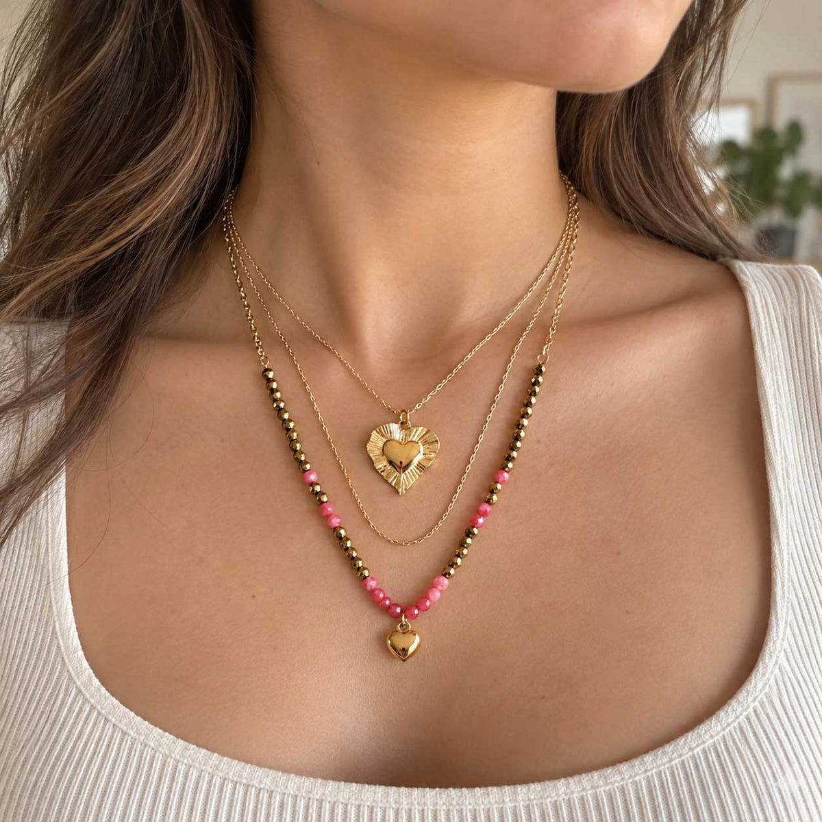 Goldene dreireihige Halskette aus Edelstahl am Hals getragen, mit facettierten Elementen und korallfarbenen Herzen als femininer Layering-Schmuck
[AI]
