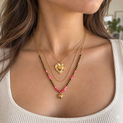 Goldene dreireihige Halskette aus Edelstahl am Hals getragen, mit facettierten Elementen und korallfarbenen Herzen als femininer Layering-Schmuck
[AI]