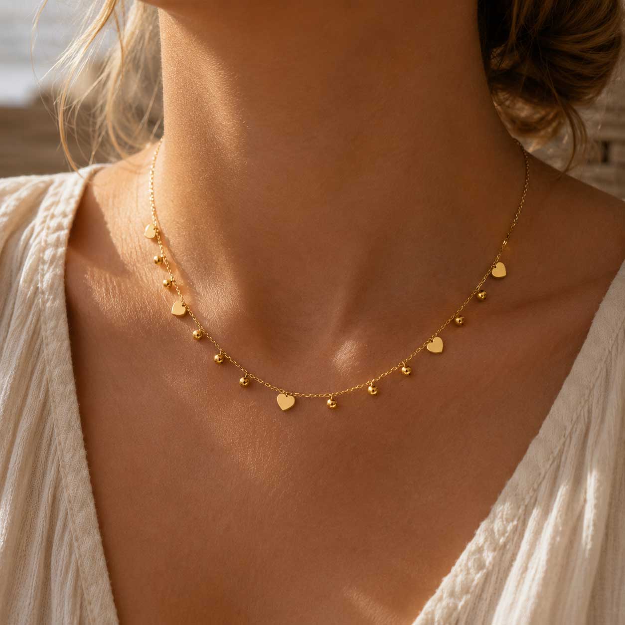 Blushheart Halskette getragen – goldene Herz-Kugel-Halskette von Saltyblush am Hals einer Frau, romantischer Look zu weißem Strickoberteil im Sonnenlicht
