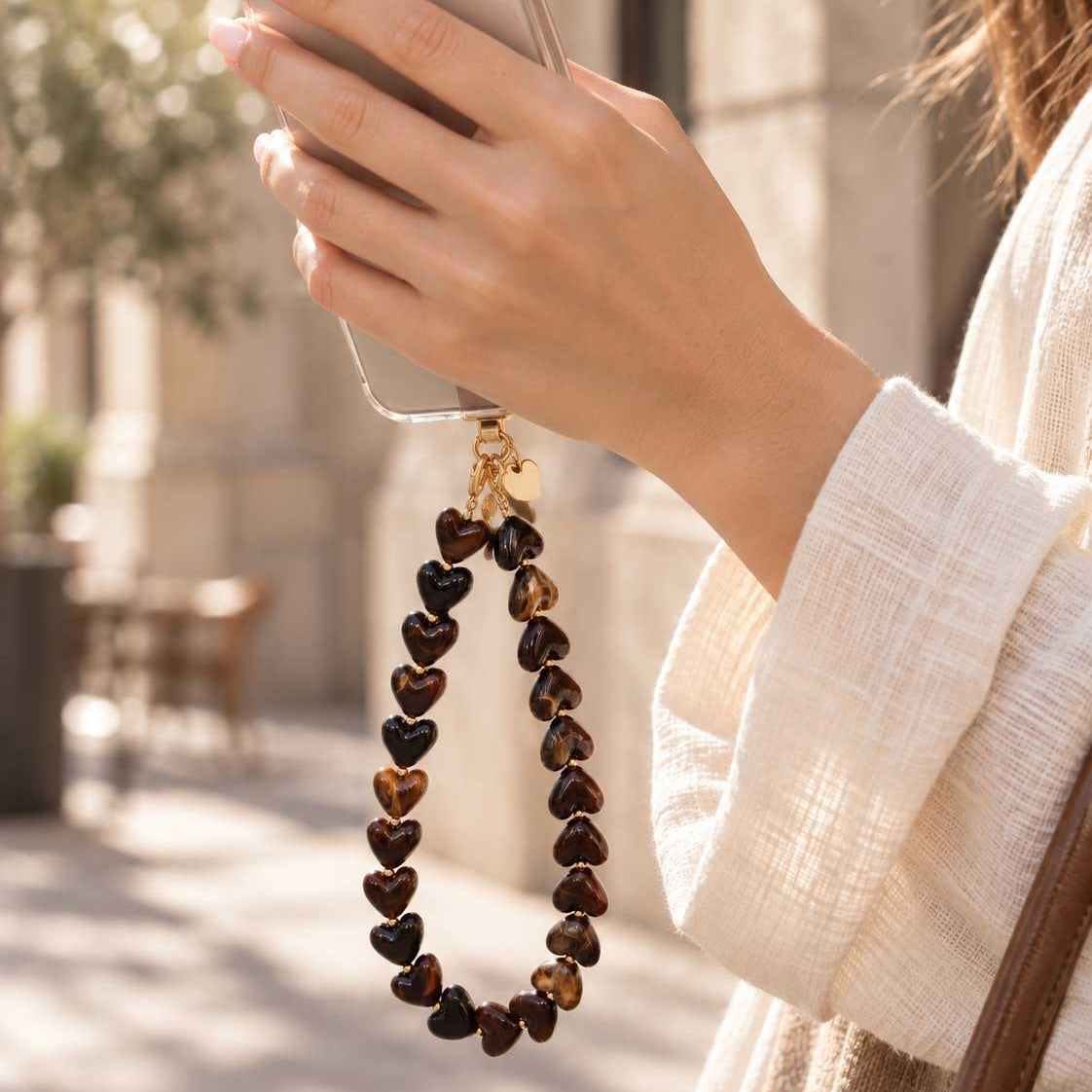 Handgefertigte braune Perlen-Handykordel am Handgelenk neben einem Smartphone, mit verspielten Herzdetails und dekorativem Phone-Charm-Design

