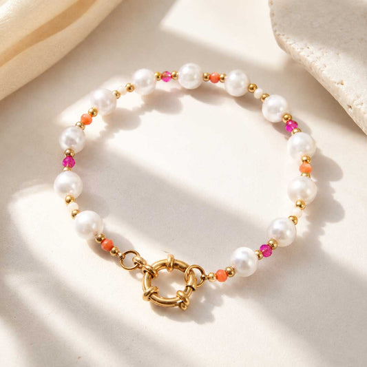 Goldenes Perlenarmband mit weißen Süßwasserperlen, pinken und orangefarbenen Kristallen sowie goldenem Ringverschluss auf cremefarbenem Untergrund mit Muschel
