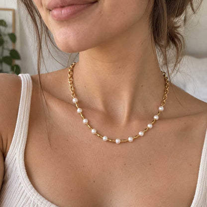 Coastal Pearl goldene Halskette getragen – brünette Frau, Nahaufnahme Hals und Dekolleté, Perlenkette mit goldenen Gliedern und Zylinder-Spacern, weißes Rippenstrick-Top, heller Innenraum
[AI]