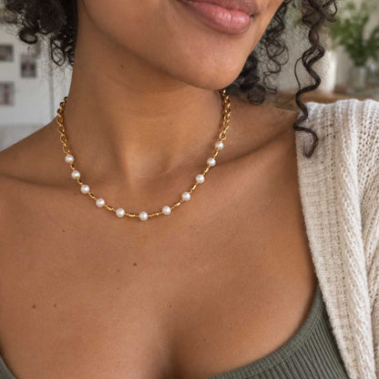 Coastal Pearl goldene Halskette Produktfoto – vollständige Kette aus O-Ring-Gliedern, weißen Perlen und goldenen Zylinder-Spacern mit Karabinerverschluss und Verlängerungskette, auf Travertin-Stein mit Muscheln und Schleierkraut
[AI]