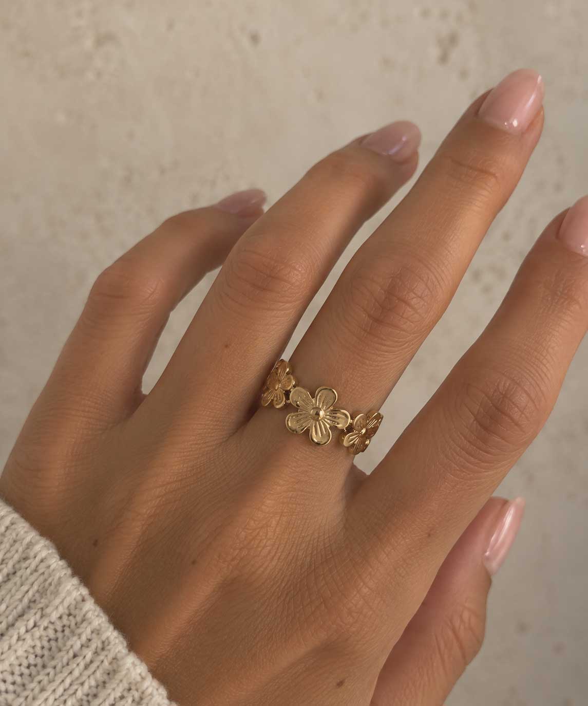 Daisy Kiss goldener Ring getragen – Nahaufnahme Frauenhand mit nude Nägeln, offener Blumenring aus vergoldetem Edelstahl am Mittelfinger, heller Hintergrund
[AI]
