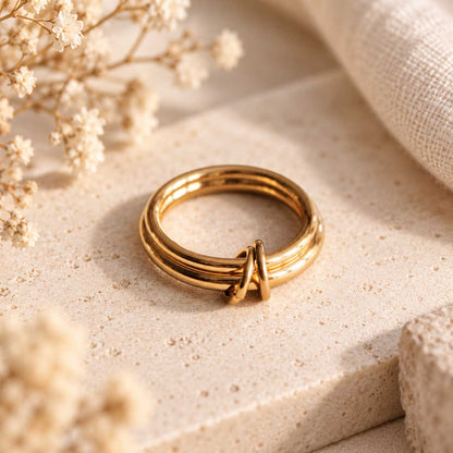 Double Loop goldener Ring – vergoldeter Edelstahlring mit zwei parallelen Bändern und Knotendetail in der Mitte, auf Travertin-Stein mit Schleierkraut, warmes Licht

