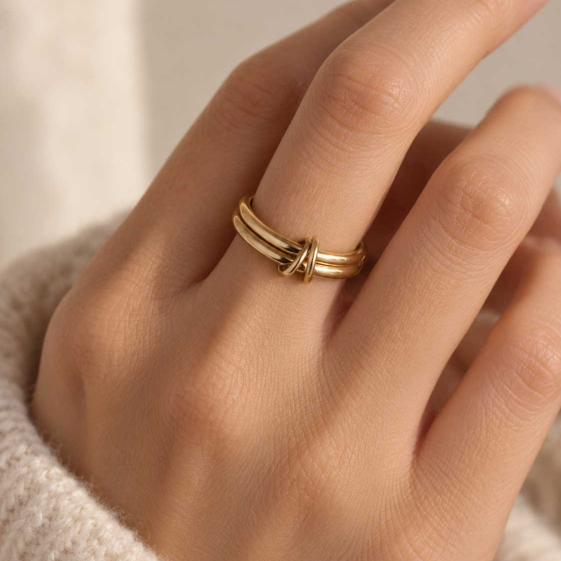 Double Loop goldener Ring getragen – Nahaufnahme Frauenhand, vergoldeter Doppelband-Ring mit Knoten am Mittelfinger, weißer Strickpullover, cremefarbener Hintergrund
[AI]