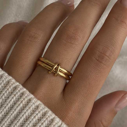 Double Loop goldener Ring getragen – Nahaufnahme Frauenhand mit nude Nägeln, vergoldeter Doppelband-Ring am Ringfinger, cremefarbener Strickpullover
[AI]