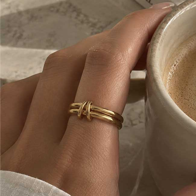 Double Loop goldener Ring getragen – Frauenhand hält cremefarbene Kaffeetasse, vergoldeter Doppelband-Ring mit Knoten am Zeigefinger, weißes Leinenoberteil, Buch im Hintergrund
[AI]