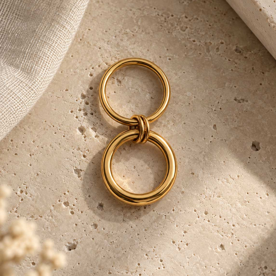 Double Loop goldener Ring Produktdetail – zwei separate vergoldete Edelstahlringe mit Knotenverbindung in der Mitte, auf Travertin-Stein, helles Sonnenlicht
