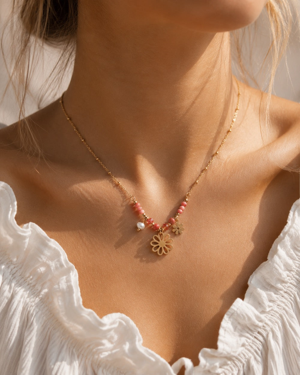 Goldene Flower Power Halskette am Hals getragen mit rosa Perlen, Süßwasserperle und zwei goldenen Blumen-Charms zu weißem Rüschenoberteil im Sonnenlicht, vergoldete Edelstahl-Satellitenkette
